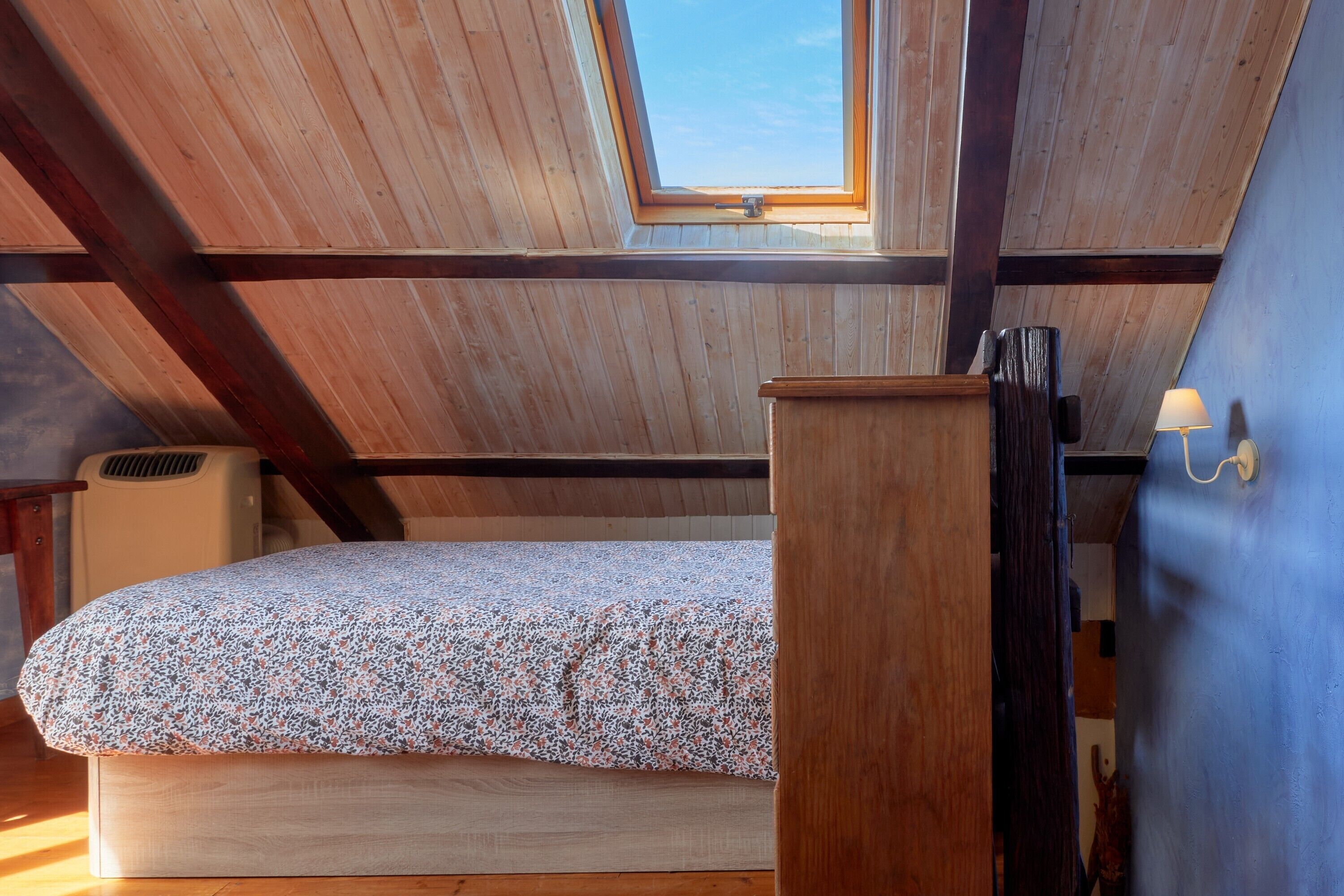 A room with a sloped ceiling, a skylight, a bed with a patterned bedspread, a bedside table, and a wall-mounted lamp.