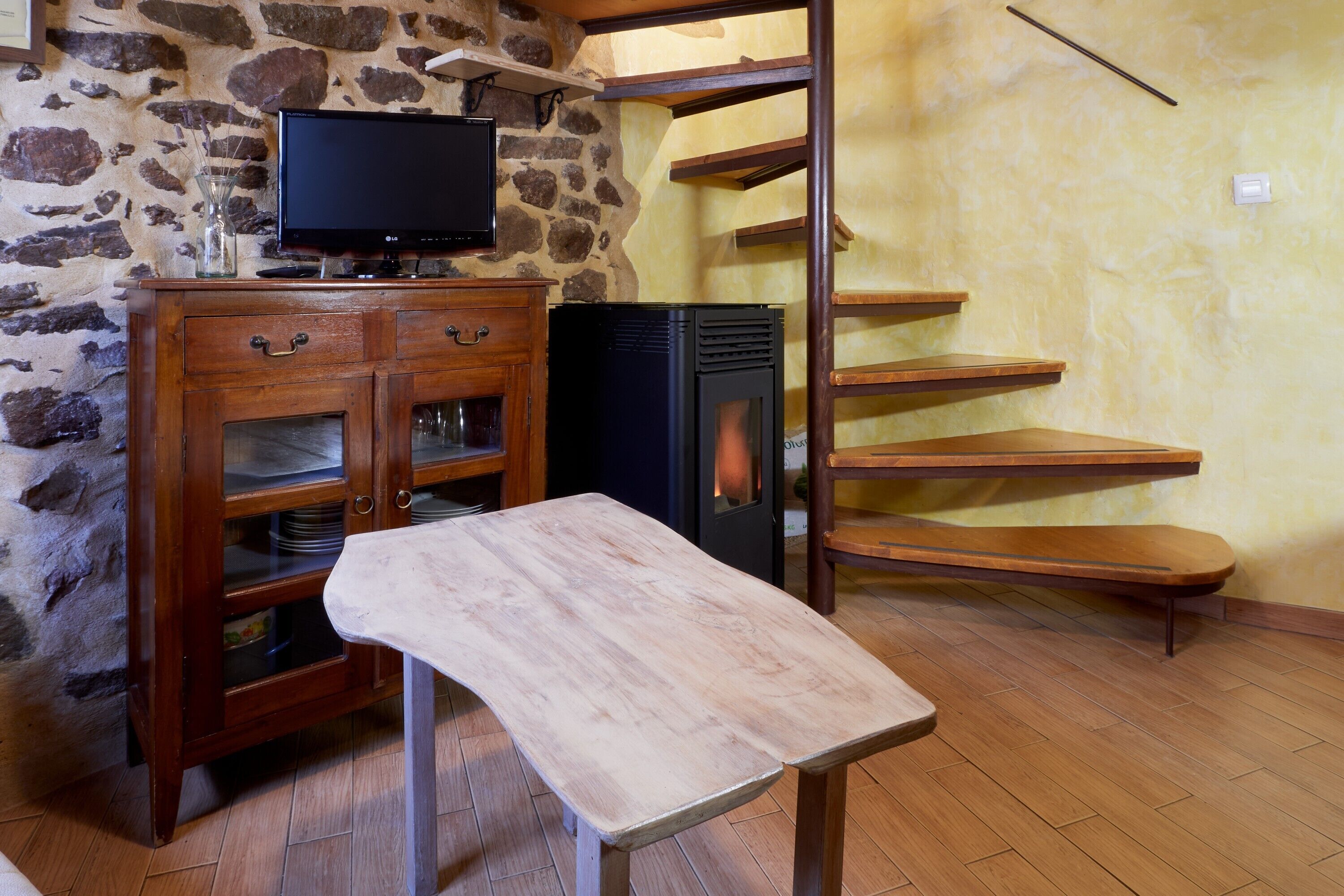 Habitació amb paret de pedra, un armari de fusta amb portes de vidre, televisió de pantalla plana, una llar de foc i una banca de fusta.