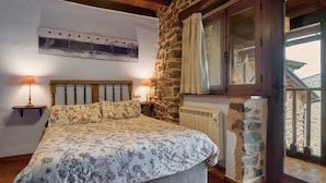 Een slaapkamer met een bed, twee bedlampen, een stenen muur, een raam met gordijnen en uitzicht op buiten.