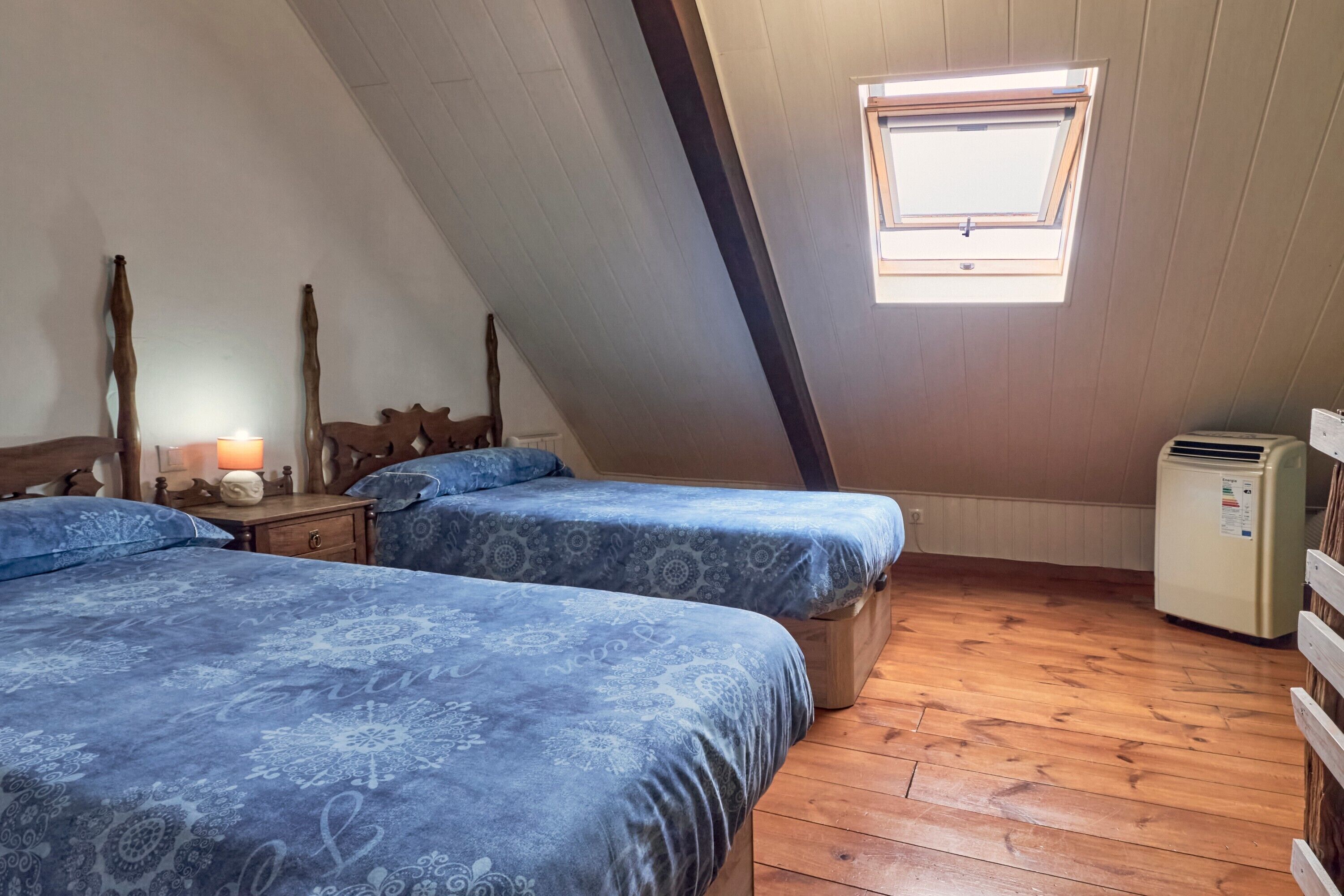 Una habitación con dos camas, una mesita con una lámpara, una ventana y un refrigerador.