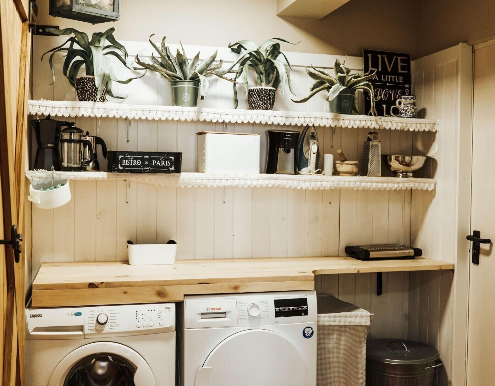 A laundry area with a washing machine, dryer, and shelves with plants and kitchen items.