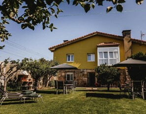 Une maison jaune avec un toit en tuiles, une cheminée en pierre et un petit porche à l’avant, avec une table et des chaises.