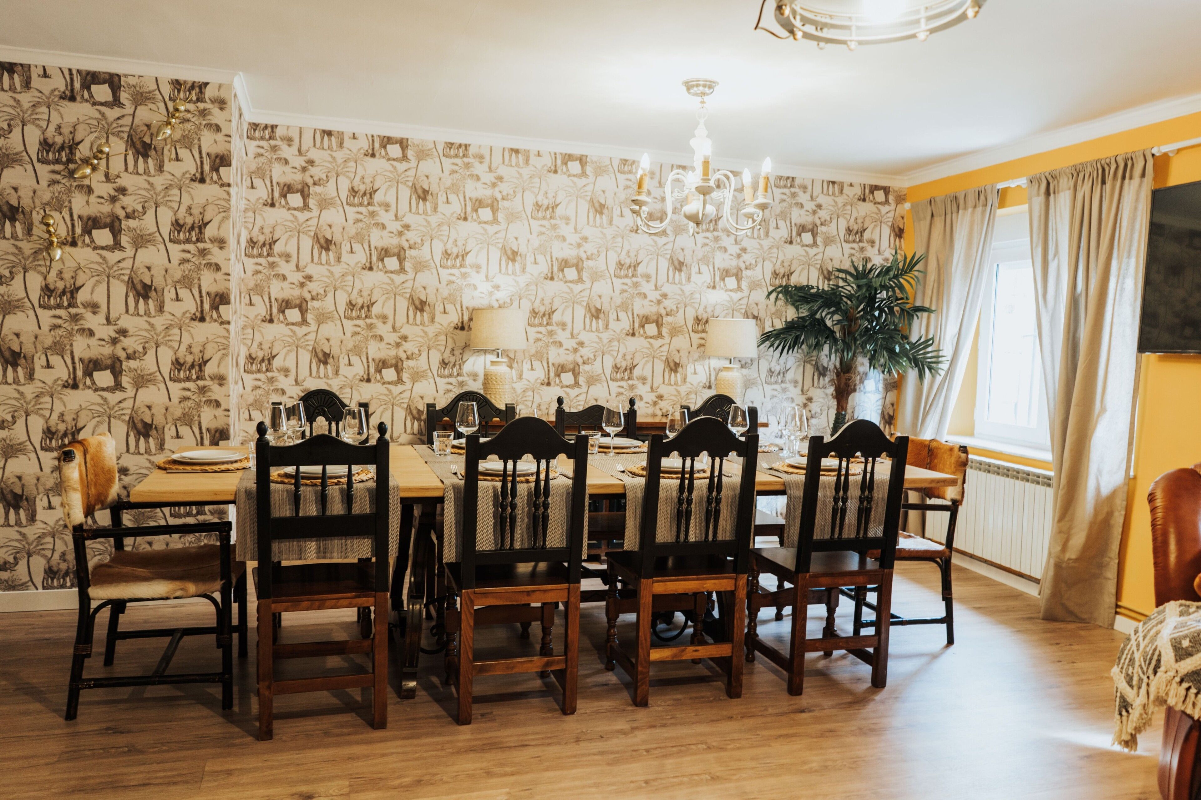 A dining room with a patterned wallpaper, wooden chairs, a chandelier, and a dining table set for six.