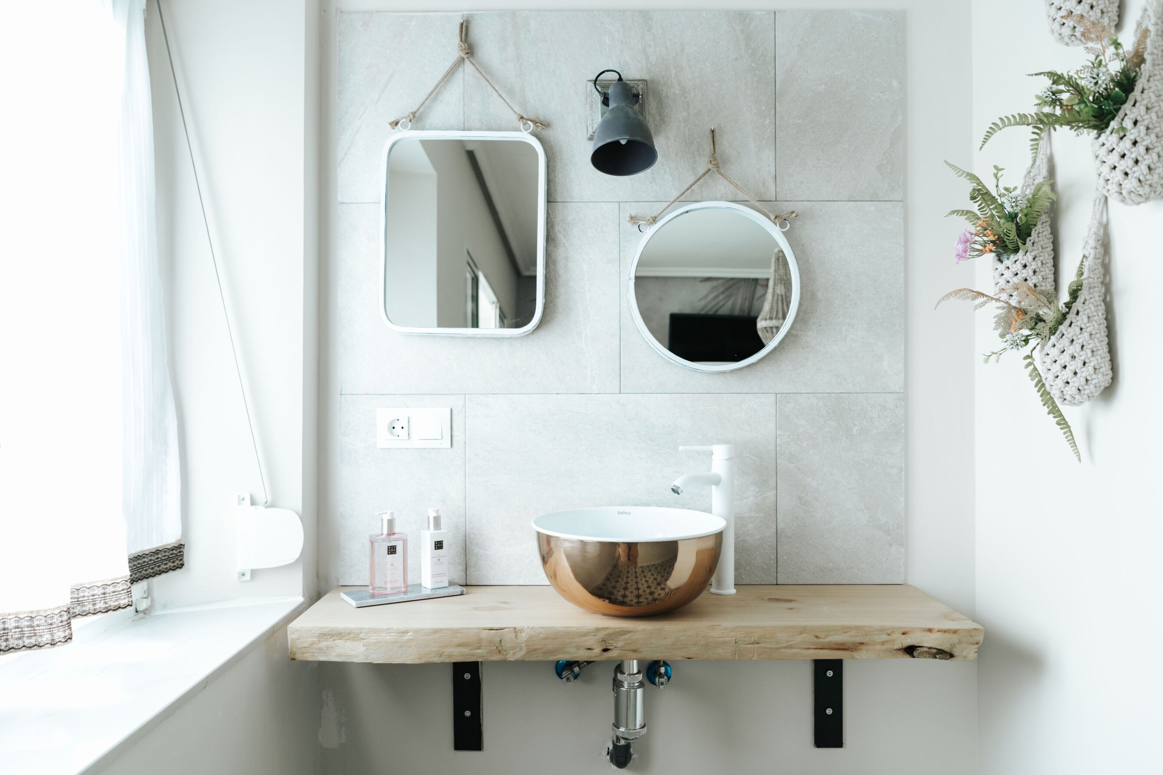A bathroom with a wooden vanity, a copper sink, two wall-mounted mirrors, a black wall-mounted light, and a window with a white curtain.