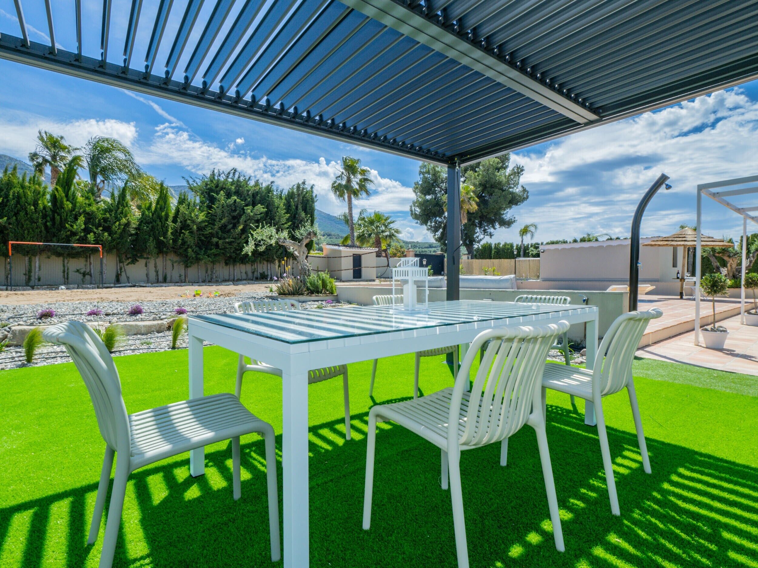 An outdoor patio with a table and chairs, surrounded by greenery and a clear sky.