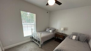 Une chambre avec deux lits, une fenêtre avec des stores, un ventilateur de plafond et une table de chevet avec une lampe.