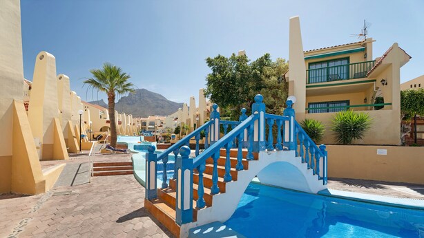 A pool area with a blue staircase leading to a pool, beige buildings, and a mountain in the background. - Vacation Apartment 'Beach Holidays' with Shared Pool, Wi-Fi and Air Conditioning (Adeje)