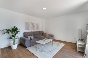 A modern living room with a grey sofa, a white coffee table, a potted plant, and a flat-screen TV on a stand.