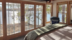 A bedroom with large sliding glass doors leading to a lake, a bed with a blanket, and a flat-screen TV mounted on the ceiling.