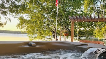 Un jacuzzi con una pérgola de madera y luces colgantes, rodeado de árboles y una bandera.