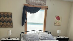A bedroom with a bed, two bedside tables, a window with blinds, a decorative sign above the window, and a wall art piece.