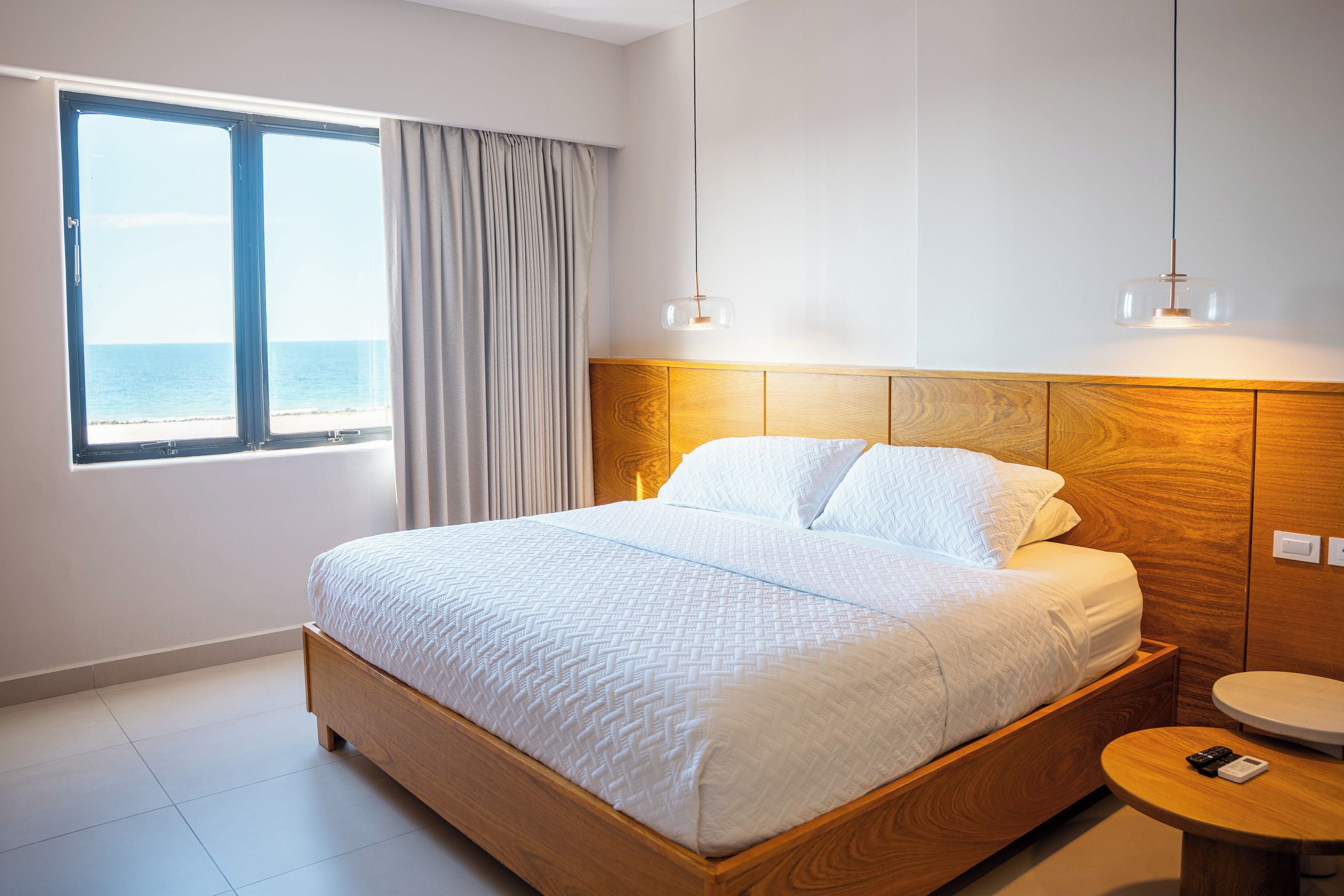 Habitación de hotel con cama, vista al mar, cabecero de madera, mesita de noche y velador.