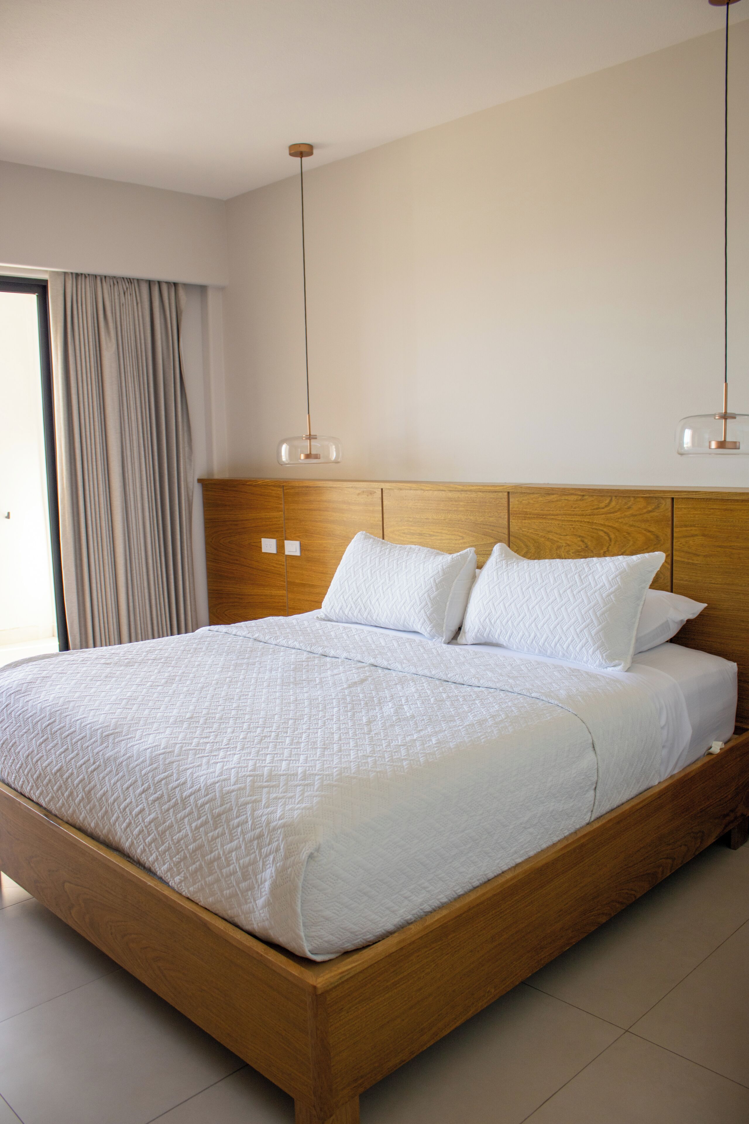 Una cama grande con cabecera de madera, ropa de cama blanca y dos lámparas colgantes.