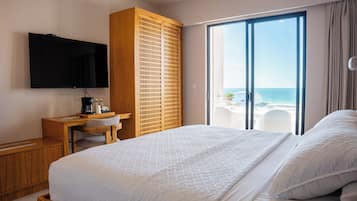 A hotel room with a bed, a desk with a coffee maker, a TV, and a view of the beach.