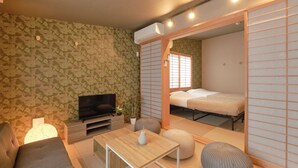 A compact living space with a sofa, TV, and a small table. A bed is visible through a sliding door, and there's a potted plant. - Ryokan Zen Asagaya by Tranova (Tokyo)
