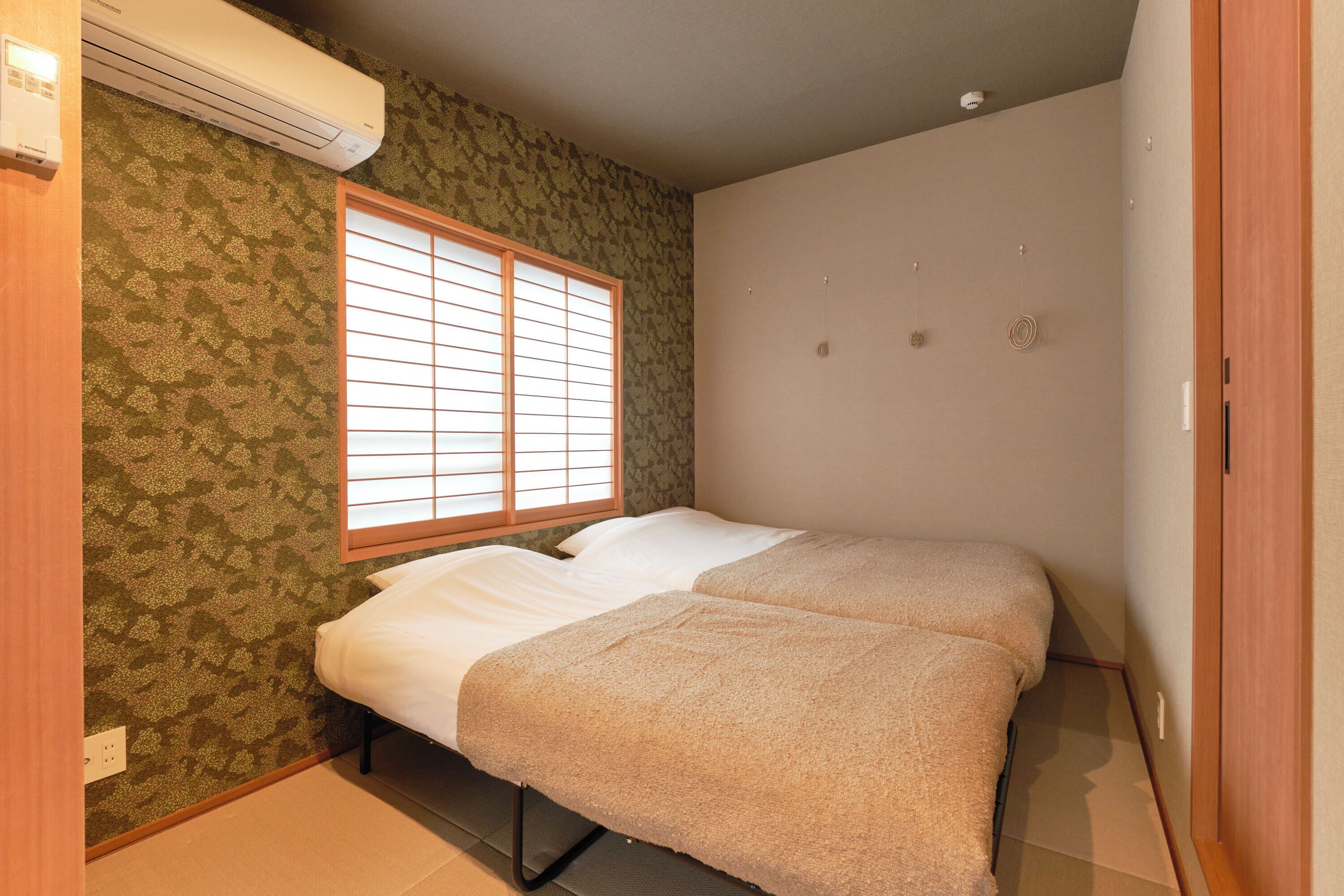 Una habitación pequeña con dos camas, una ventana con persianas de madera, un aire acondicionado montado en la pared y una pared lisa con un interruptor de luz.