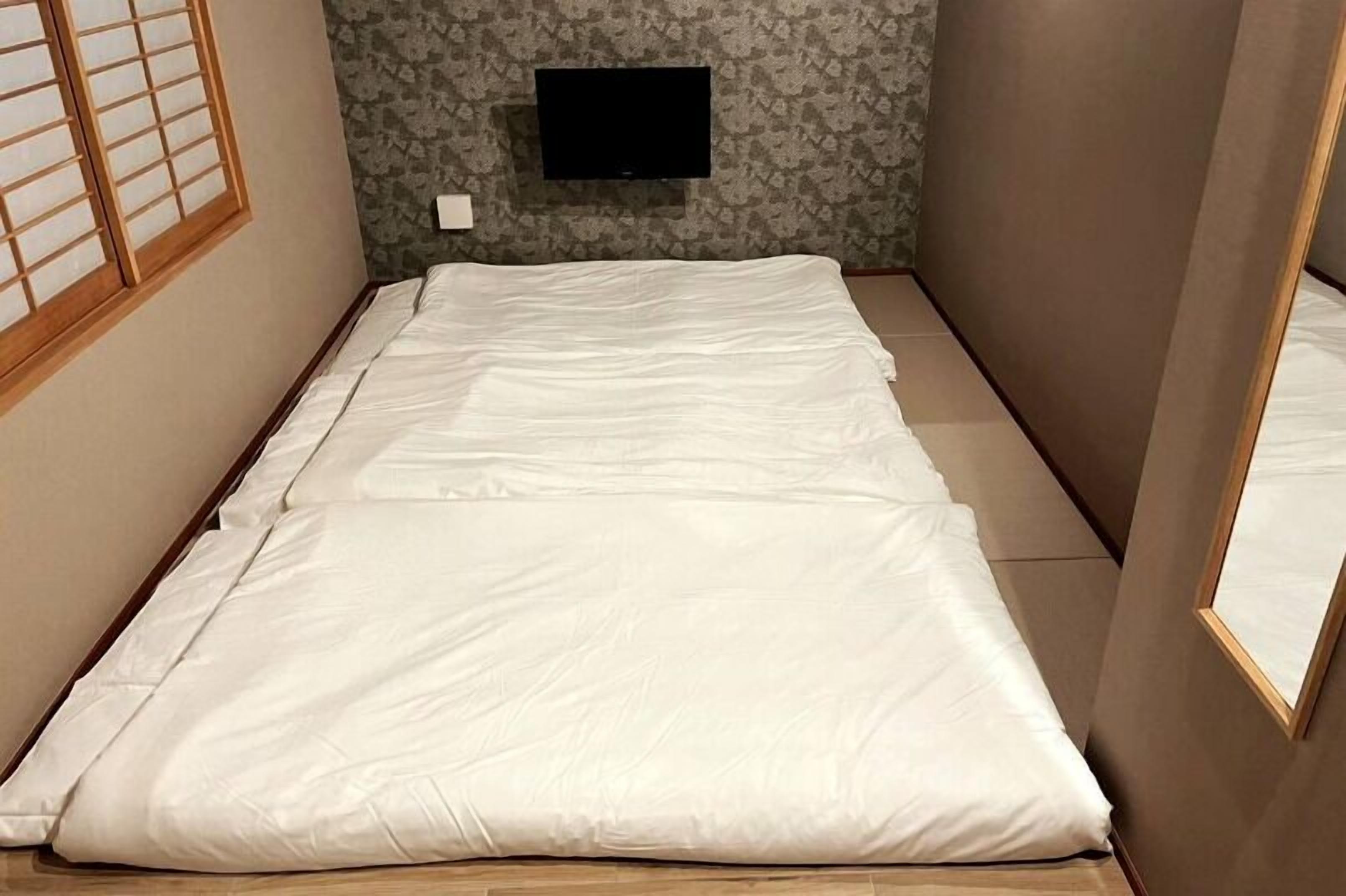 A room with a bed, a television mounted on the wall, and a window with wooden blinds.