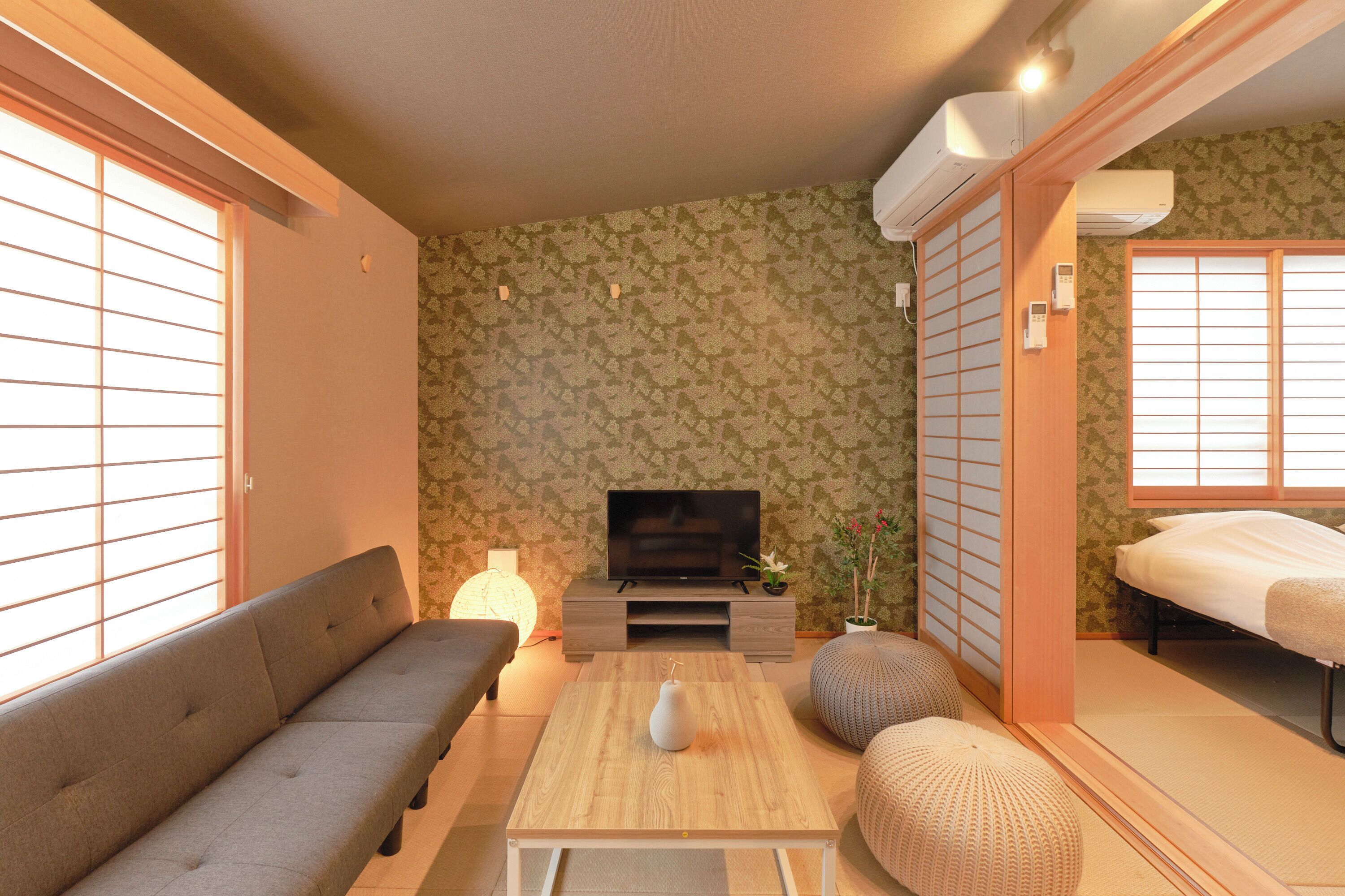 A traditional Japanese-style living room with tatami flooring, a low wooden table, a sofa, a television, and a futon bed.