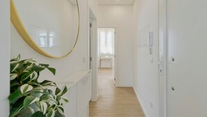 Un couloir aux murs blancs, avec un plancher en bois et un grand miroir sur le côté gauche. Il y a deux portes sur le côté droit, et une plante au premier plan.