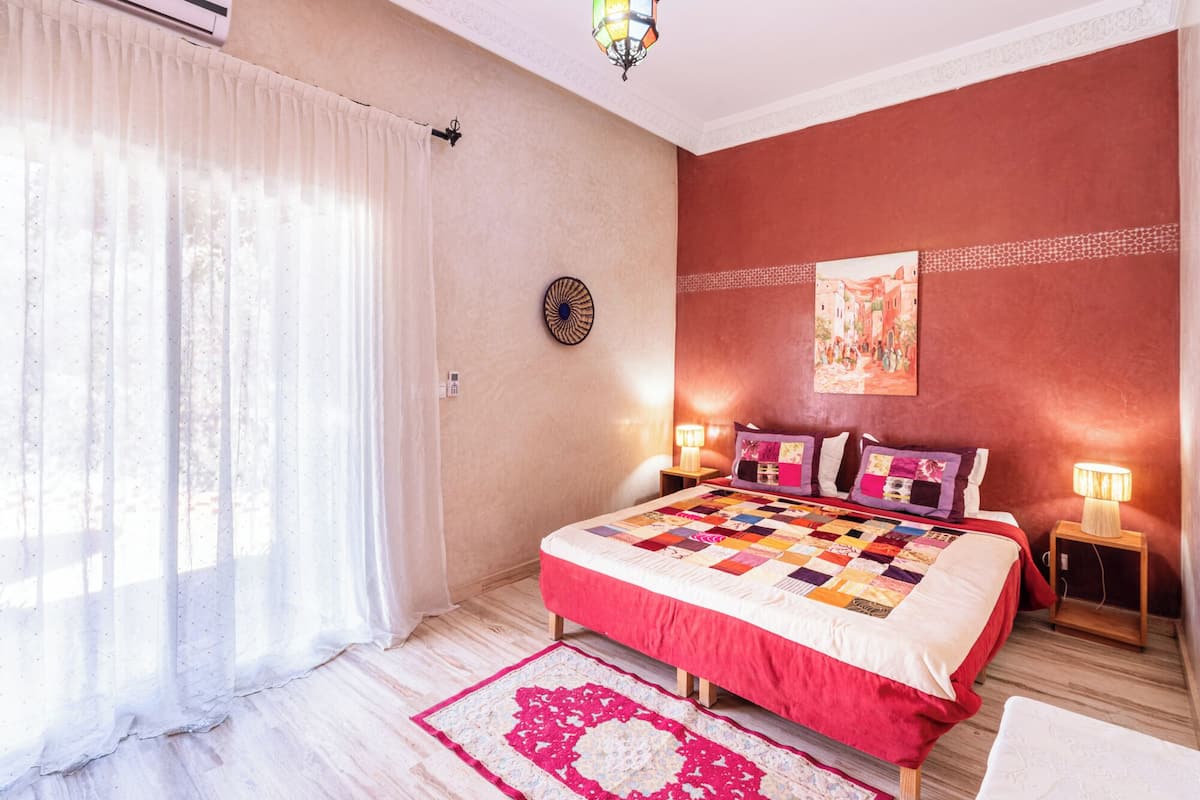 Un dormitorio con una cama grande, dos lámparas de noche, una ventana con cortinas translúcidas, un reloj de pared y una alfombra decorativa.