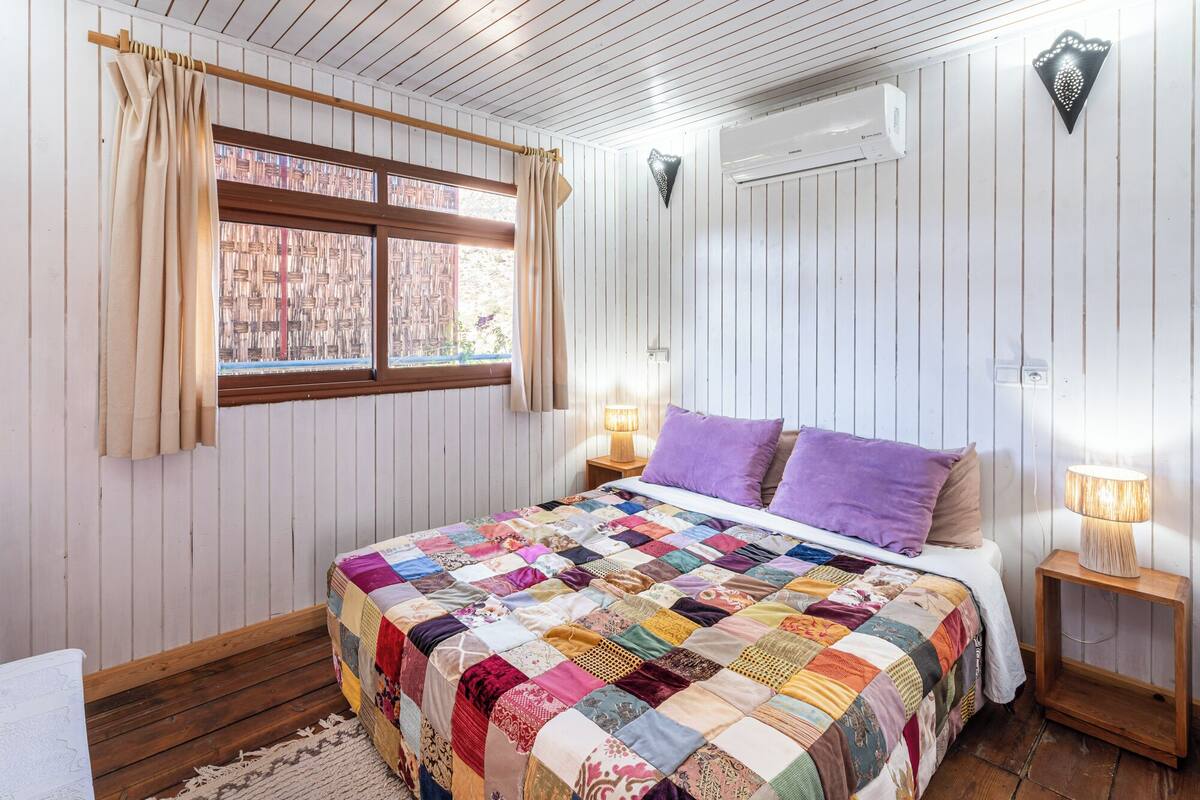Un dormitorio con suelo de madera, colcha acolchada, ventana con cortinas, unidad de aire acondicionado y mesita de noche con lámpara.