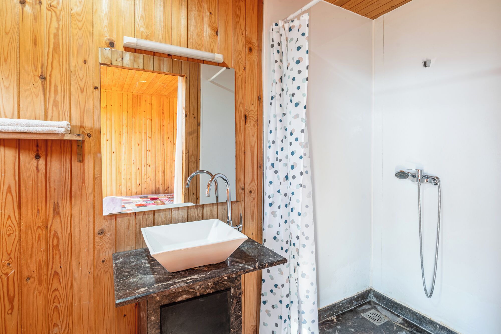 A bathroom with a wooden wall, a white sink with a marble countertop, a shower with a polka dot curtain, and a mirror above the sink.
