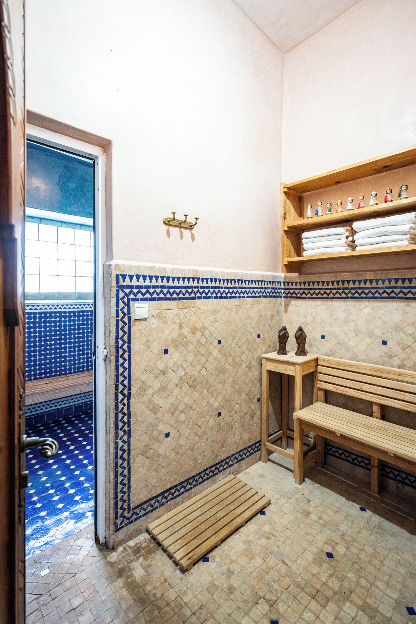 A bathroom with a tiled floor, a bench, a shelf with towels, and a door leading to a tiled room.
