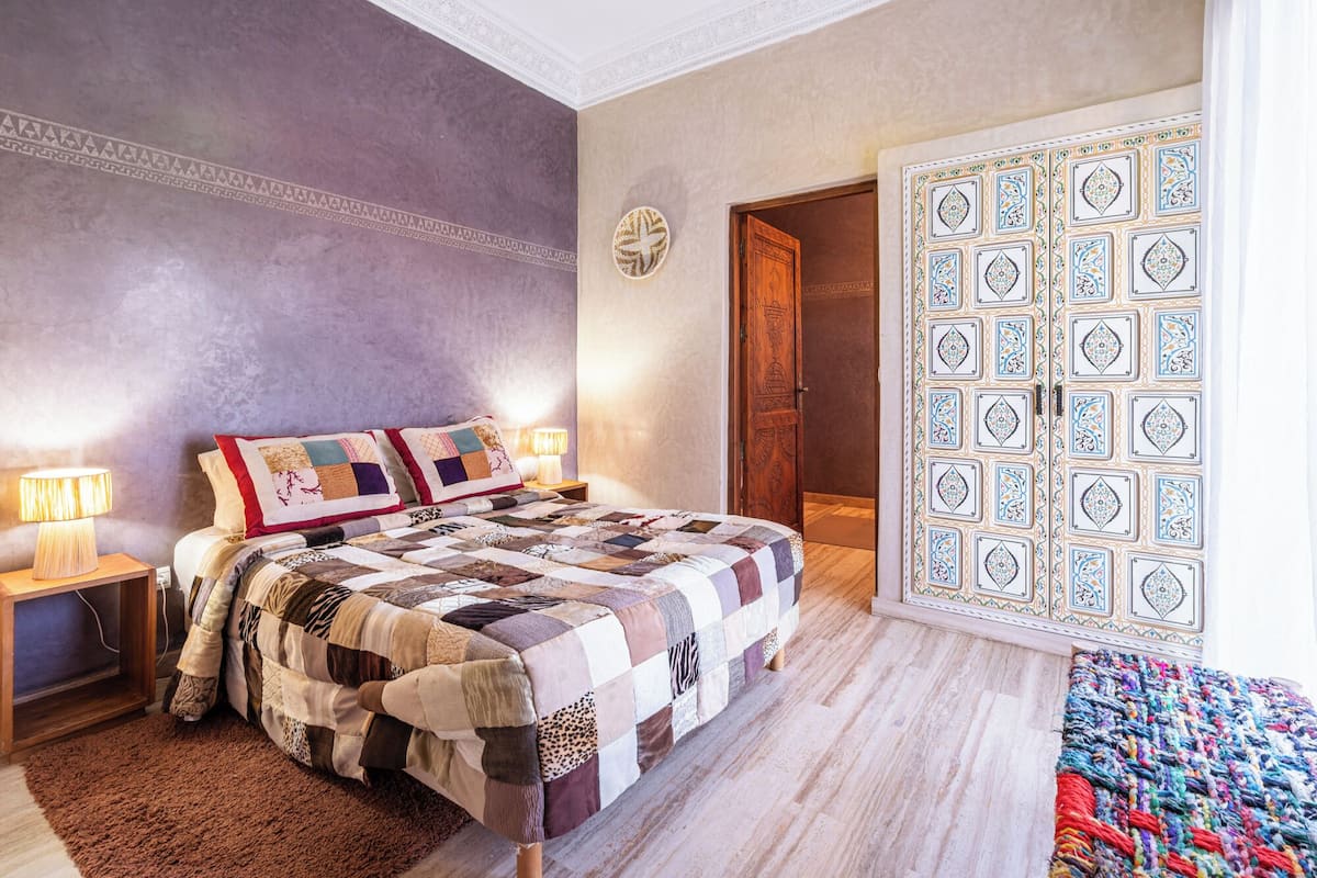Un dormitorio con una cama grande, una mesita de noche de madera, una lámpara de pared y una puerta con diseño.