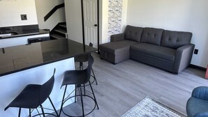 Una cocina moderna con encimera negra y dos taburetes de bar, un sofá seccional gris y una alfombra estampada.