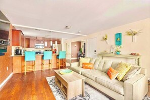 Una cocina moderna con una isla central, un comedor con sillas, un sofá con cojines y una estantería decorativa con plantas.