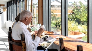 Un homme est assis à une table en bois, lisant un magazine, avec vue sur la verdure et les bâtiments environnants.