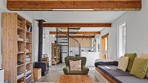 Un salon chaleureux avec une table basse en bois, un canapé avec des coussins, une bibliothèque et un escalier en colimaçon menant à la cuisine.