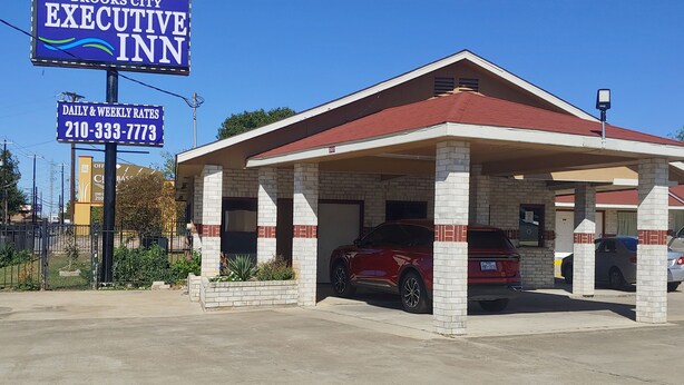 A hotel with a red roof and white pillars, a red SUV parked in front, and a sign advertising daily and weekly rates. - Brooks City Executive Inn (San Antonio)