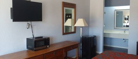 A hotel room with a wooden desk, a flat-screen TV, a microwave, a lamp, a bed with a patterned bedspread, a bathroom with a sink and mirror.