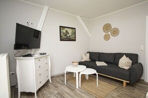 A modern living room with a flat-screen TV mounted on a white cabinet, a grey sofa with patterned cushions, a small white table, and a framed landscape painting on the wall.