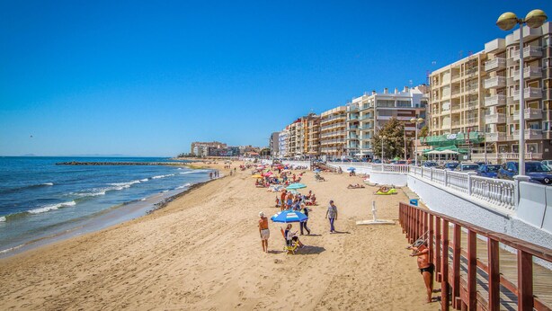A beach with a wooden promenade, beach umbrellas, and a row of multi-story buildings with balconies.