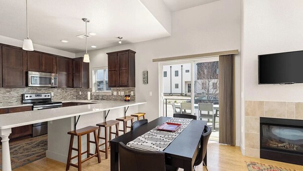 A modern kitchen with dark wood cabinets, a white countertop, and a built-in oven. There is a dining area with a black table and wooden stools. A flat-screen TV is mounted on the wall above the fireplace. - Cozy townhome in old town north dogs, Bikes. (Fort Collins)