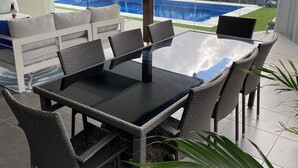 Eine Terrasse im Freien mit Tischplatte aus Glas und Korbstühlen, mit Blick auf einen Swimmingpool und einen Garten.