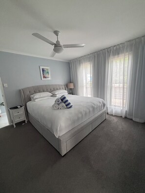 Ein Schlafzimmer mit Bett, Nachttisch, Deckenventilator, einem Fenster mit durchsichtigen Vorhängen und einem Wandbild.