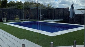 Ein rechteckiger Pool mit Glasveranda, umgeben von einer Holzterrasse und einer Rasenfläche.