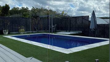 A rectangular swimming pool with a glass enclosure, surrounded by a wooden deck and a grassy area.
