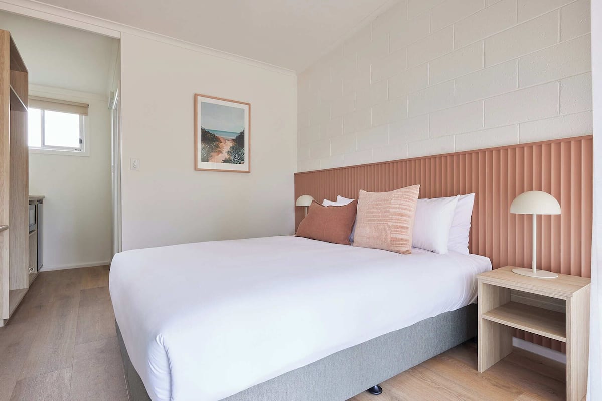 A bedroom with a bed, a nightstand, a lamp, a picture on the wall, and a window.
