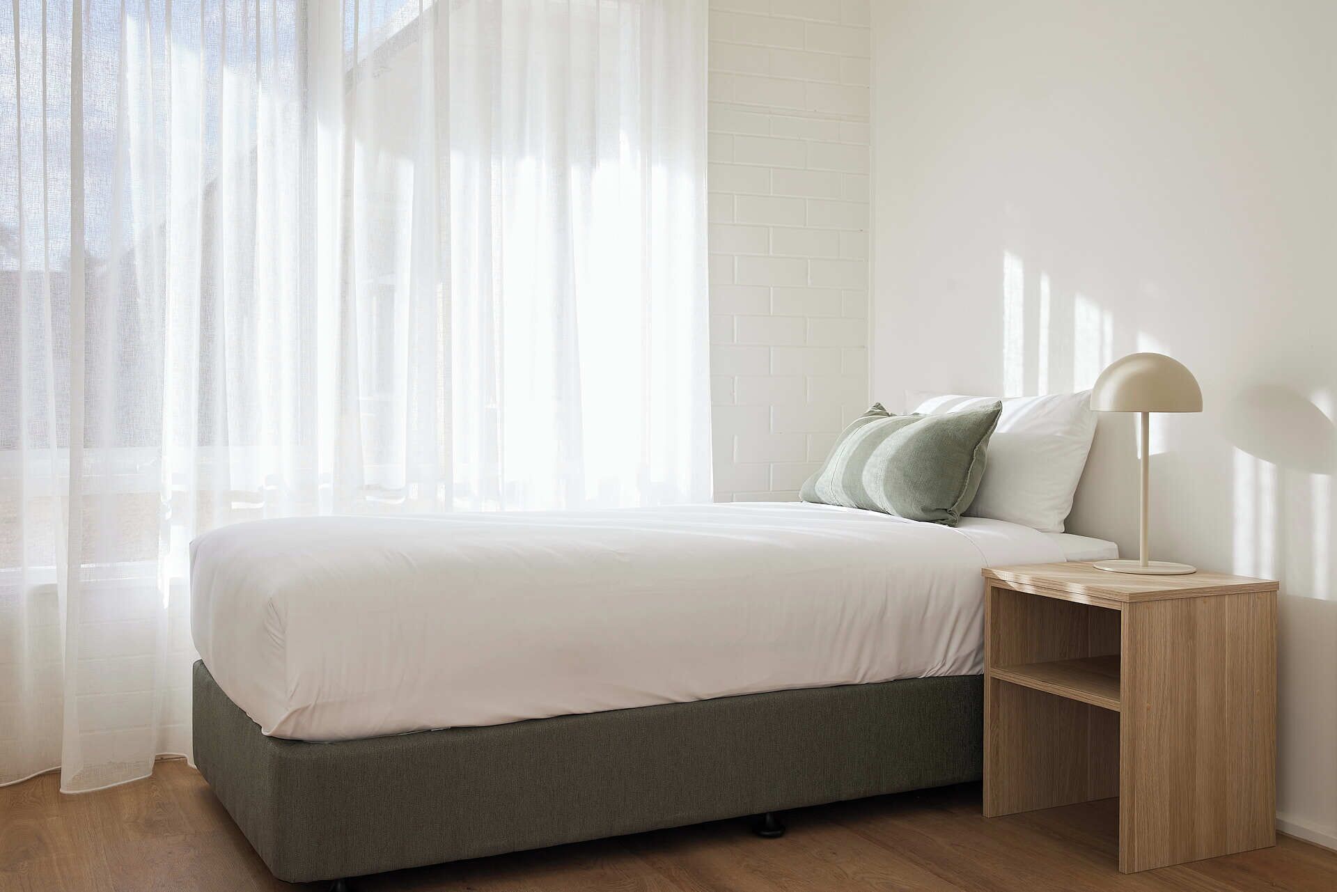 A bedroom with a bed, a nightstand, a lamp, and sheer curtains.