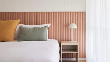 A neatly made bed with two pillows, a bedside table with a lamp, and a wall with vertical blinds.