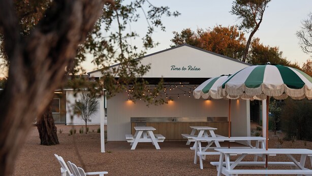 Un'area patio con tavoli e sedie da picnic bianche, un ombrellone a righe verdi e bianche e un edificio con il cartello 'Time to Relax' sullo sfondo.