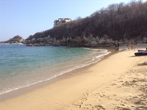 A sandy beach with clear water, rocky cliffs, and a building on the hillside.