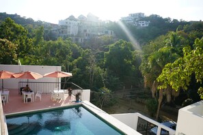 A swimming pool with white chairs and umbrellas, surrounded by lush greenery and overlooking a hillside with buildings. - Casa Esperanza (San Pedro Pochutla)