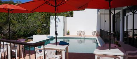 Zona junto a la piscina con sillas blancas y sombrillas rojas, una piscina pequeña y un edificio blanco al fondo.