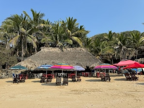A beach with thatched-roof huts, colorful umbrellas, and wooden lounge chairs.
