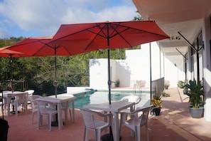 Un patio al aire libre con mesas y sillas de plástico blanco, un paraguas rojo y una piscina.